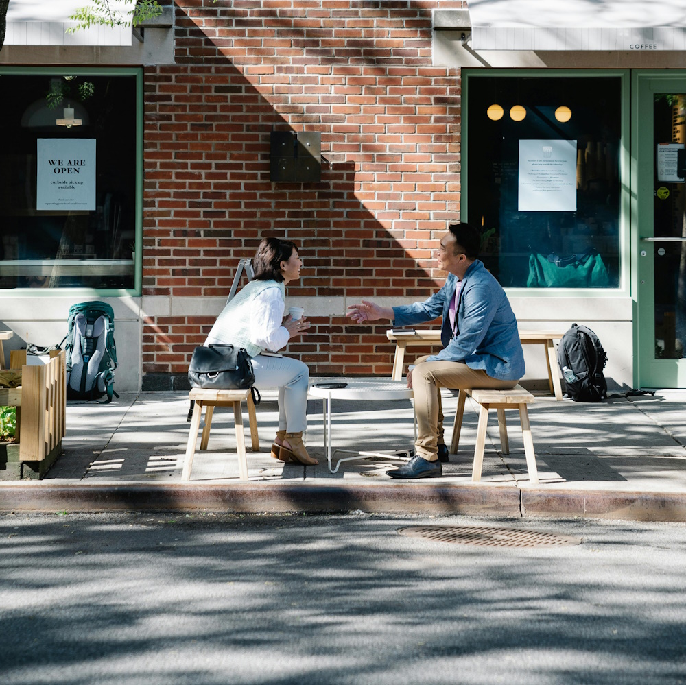 2 people sitting on benches facing one another on the sidewalk outside a business.